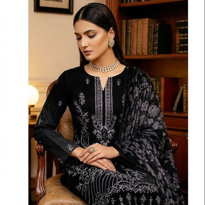 Woman in black embroidered outfit sitting in a room with wooden furniture and decor.