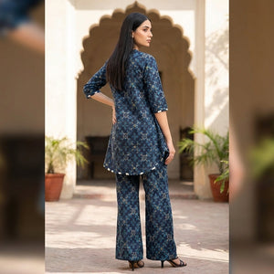 Woman in a blue patterned outfit standing in a sunlit outdoor setting with arches and plants.