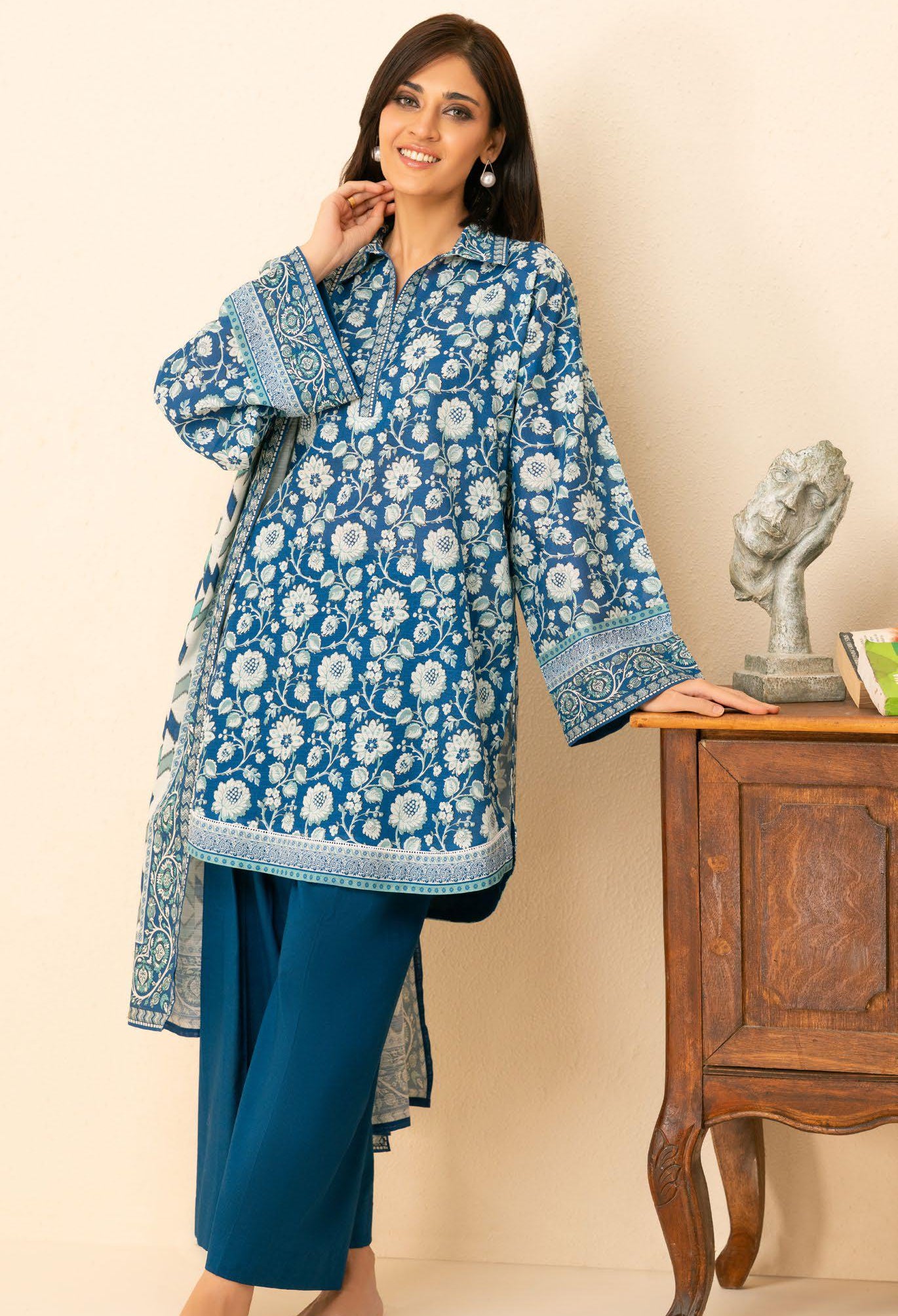 Woman wearing a blue floral traditional outfit standing next to a wooden cabinet.