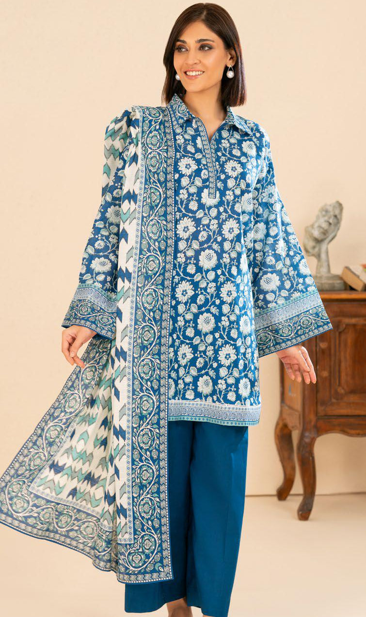 Woman wearing a blue floral traditional outfit in a room with a beige wall and wooden furniture.