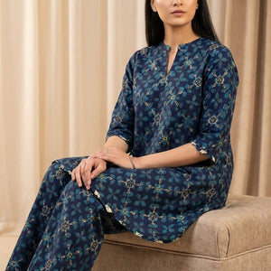 Woman wearing a blue floral dress sitting on a beige ottoman with beige curtains in the background